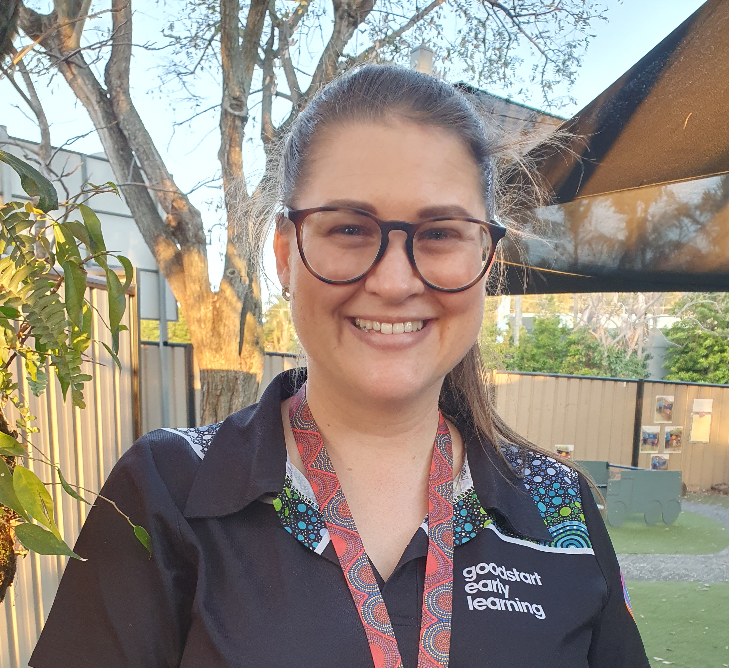 A woman who's a teacher standing in a playground smiling. She's wearing a black shirt and she has glasses.